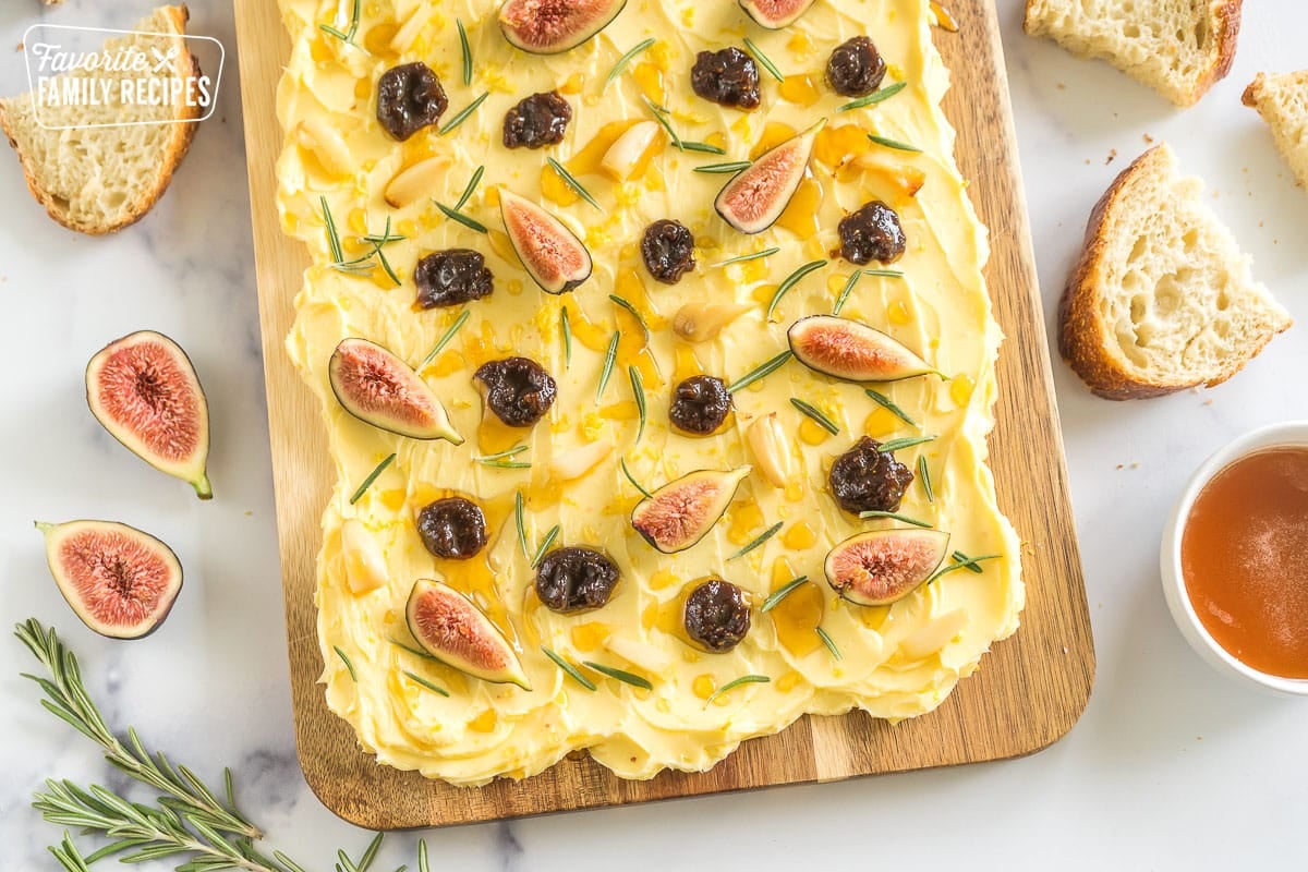 A butter board surrounded by bread slices, rosemary, and a bowl of honey.