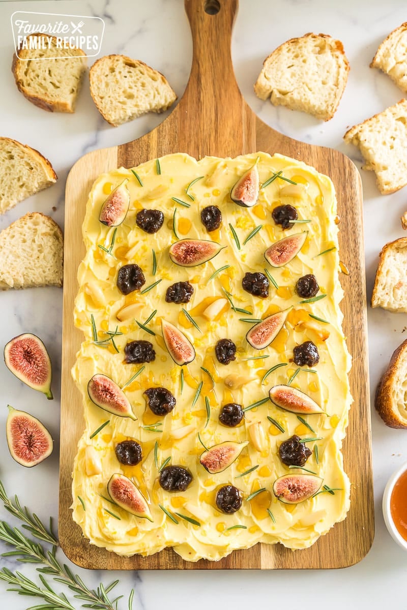 A butter board surrounded by bread slices, rosemary, and a bowl of honey.