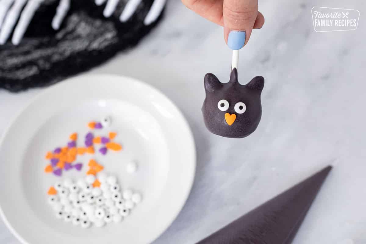 Placing eyes and heart nose onto black cat Halloween cake pop.