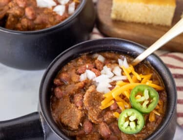 Steak chili with cheese, onion and jalapeños in black bowls with spoons and cornbread.