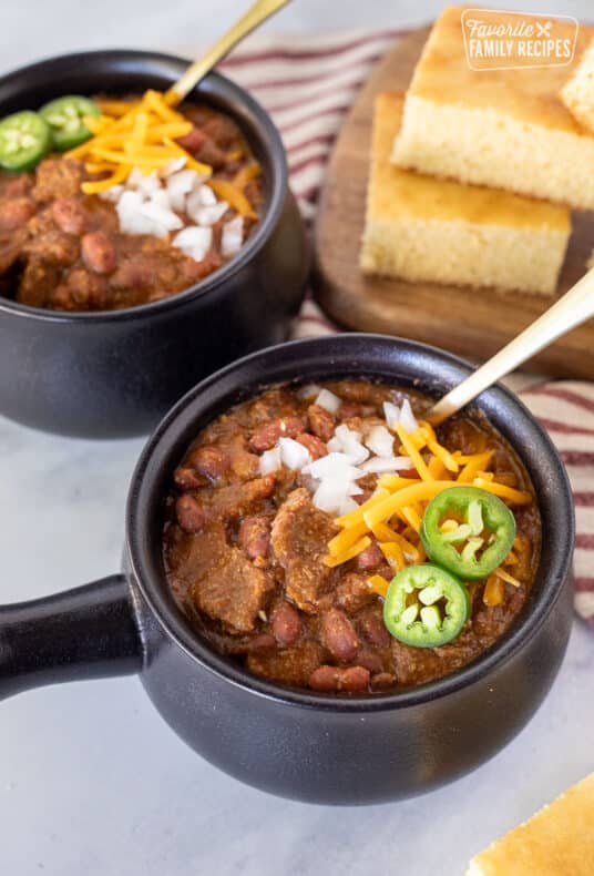 Steak chili with cheese, onion and jalapeños in black bowls with spoons and cornbread.