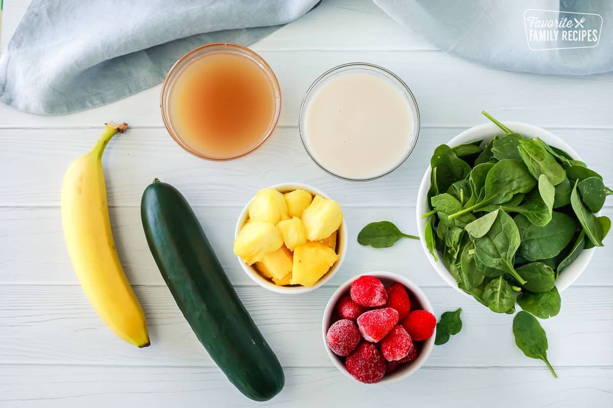 Ingredients to make a cucumber smoothie, including banana, cucumber, pineapple, frozen strawberries, apple juice, almond milk, and spinach.