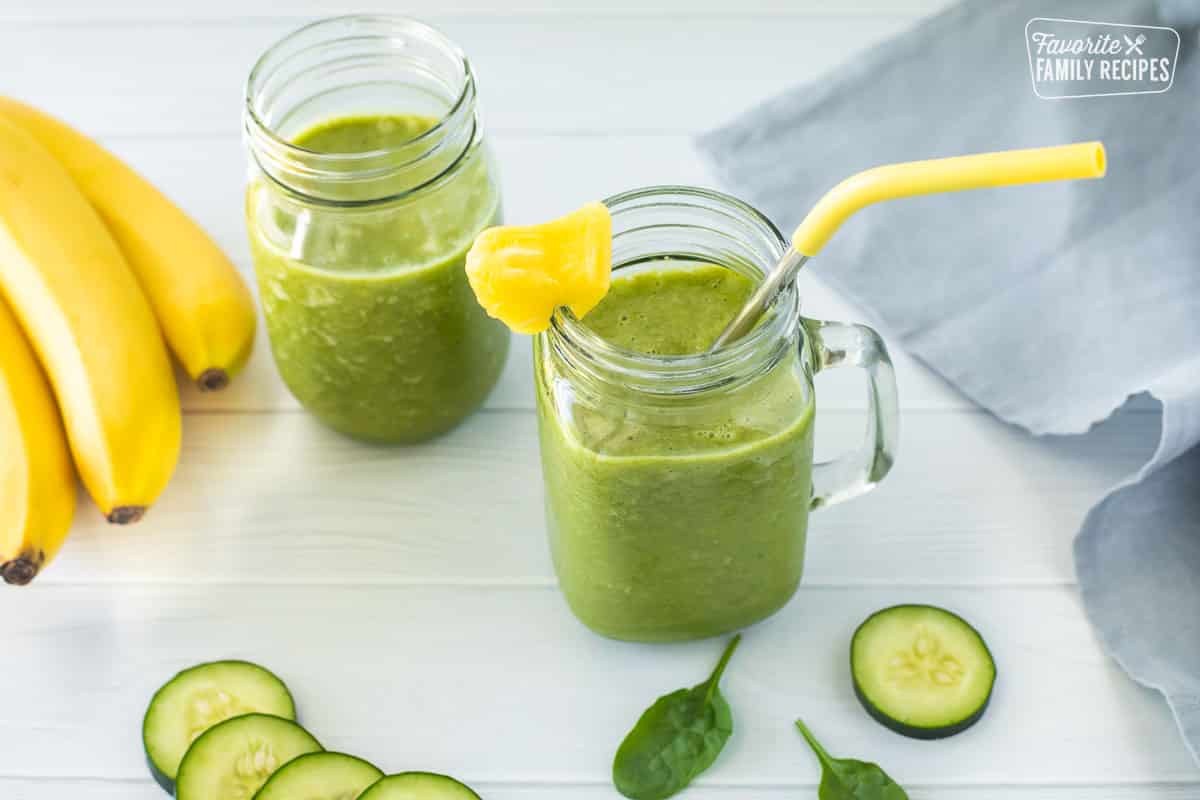 Overhead shot of two jars filled with cucumber smoothie, with the first jar having a straw and a pineapple on the rim. Bananas and cucumbers are also seen in the background.