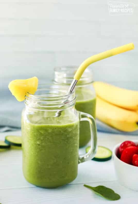 Cucumber smoothie inside a jar with a straw and a slice of pineapple on the rim. Ingredients in the background including bananas, cucumbers and strawberries.