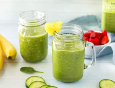 Two green smoothies in jars and a blender filled with green smoothie are seen. Ingredients including strawberries, bananas and cucumbers are featured as well.