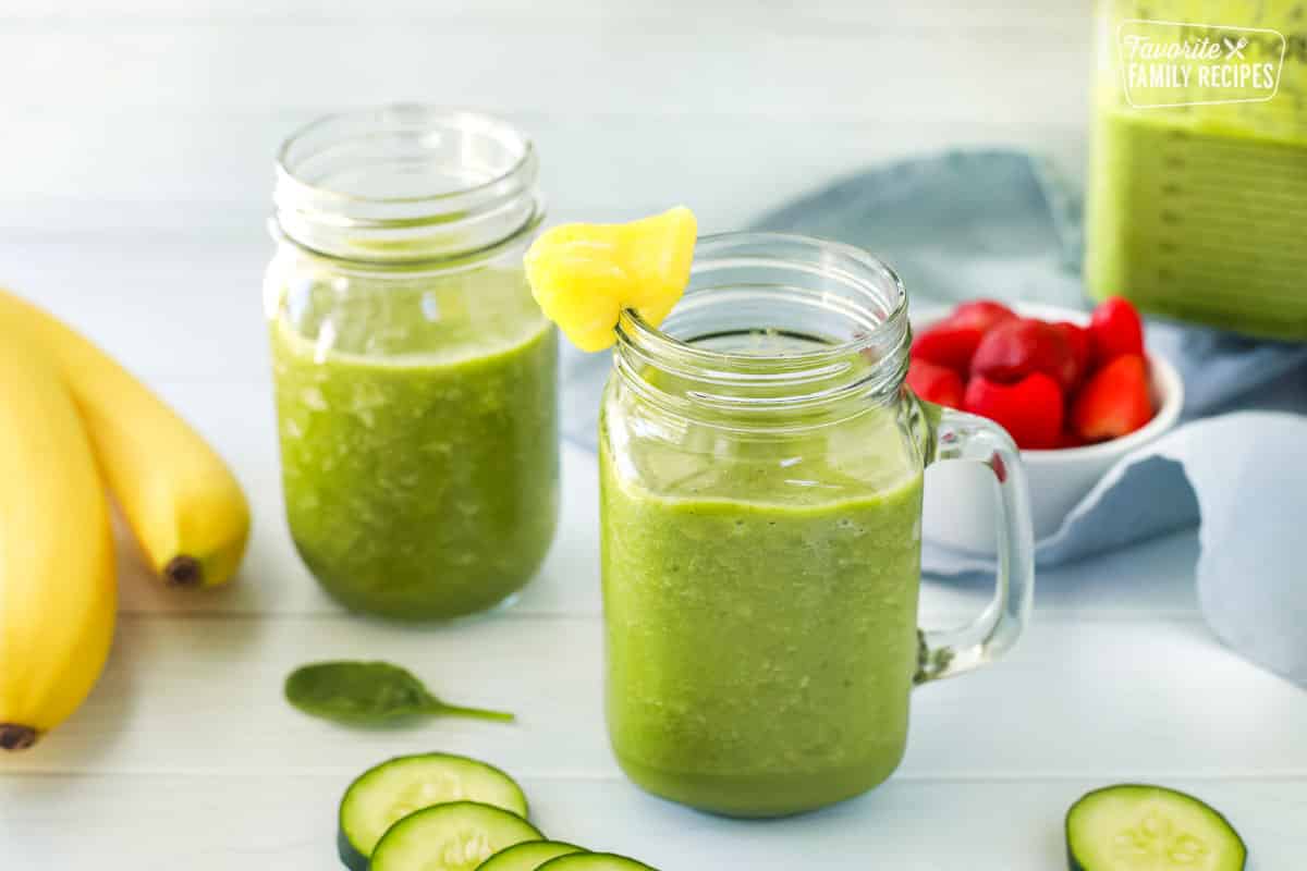 Two cucumber smoothies in jars and a blender filled with green smoothie are seen. Ingredients including strawberries, bananas and cucumbers are featured as well.