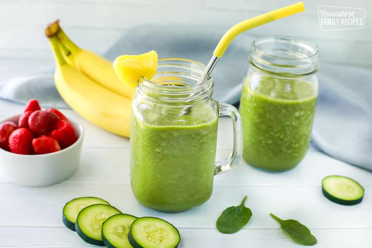 Green smoothie inside a jar with a straw and a slice of pineapple on the rim, a second jar of the smoothie is also behind it. Ingredients including strawberries, bananas and cucumbers are also seen.