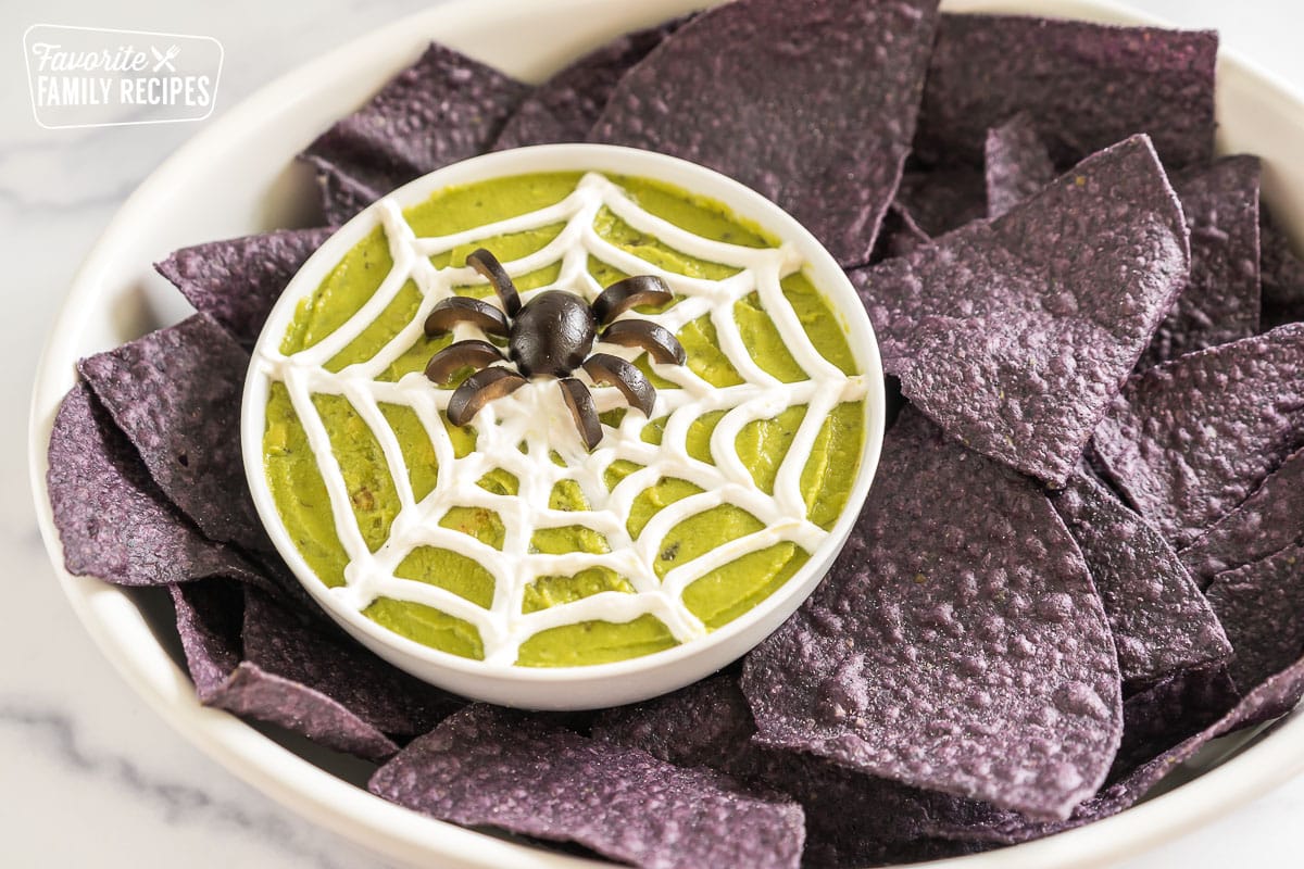 a bowl of guacamole with a sour cream spider web and an olive spider with blue corn tortilla chips
