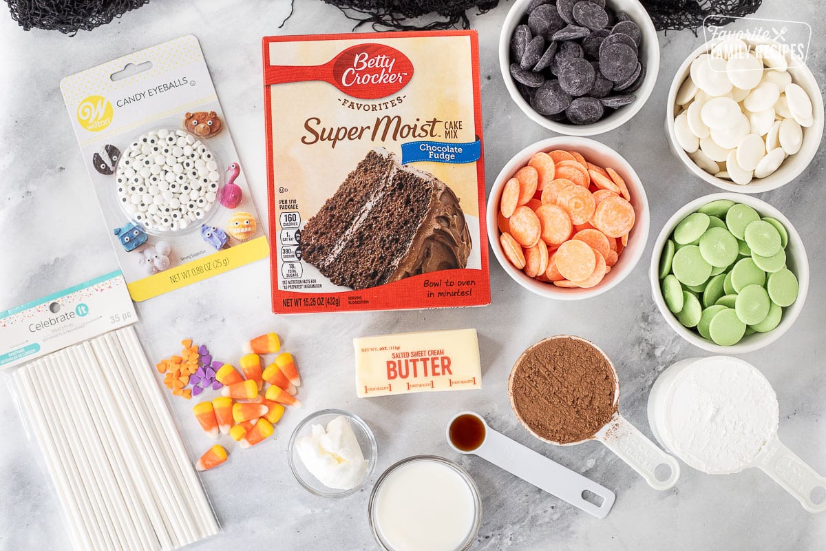 Ingredients to make Halloween Cake Pops including cake mix, butter, powdered sugar, cocoa powder, vanilla, milk, shortening, candy eyes, paper sticks, candy corn, heart sprinkles and chocolate melts in green, white, black and orange.
