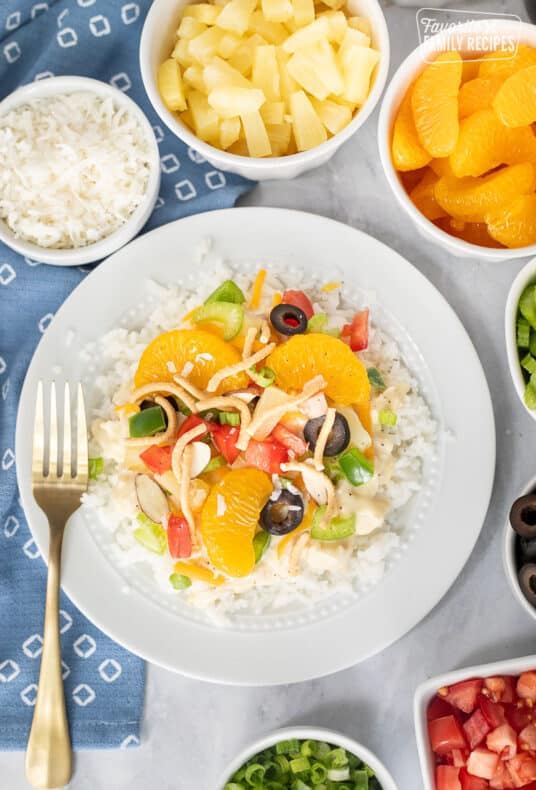 Hawaiian Haystack on a plate topped with green onion, cheese, rice noodles, green peppers, almond slices, tomatoes, celery, black olives, coconut, pineapple and mandarin oranges.