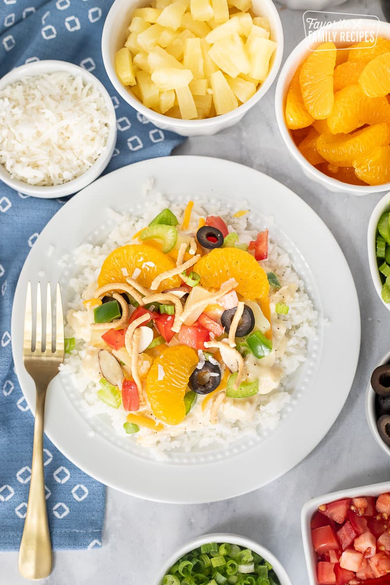 Hawaiian Haystack on a plate topped with green onion, cheese, rice noodles, green peppers, almond slices, tomatoes, celery, black olives, coconut, pineapple and mandarin oranges.