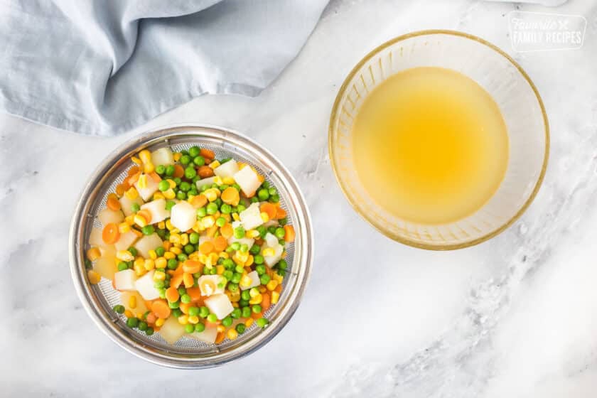 Colander of peas, carrots, corn and potatoes next to a bowl of chicken broth for Homemade Chicken Pot Pie.