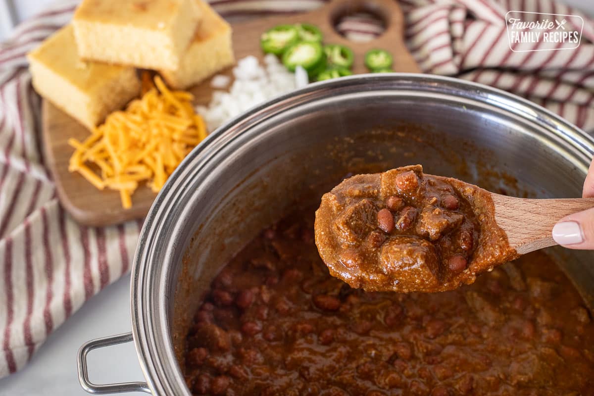 Scooping up a serving of Texas Chili in a wooden spoon. Garnishes in the background of cornbread, cheese, onion and jalapeño.