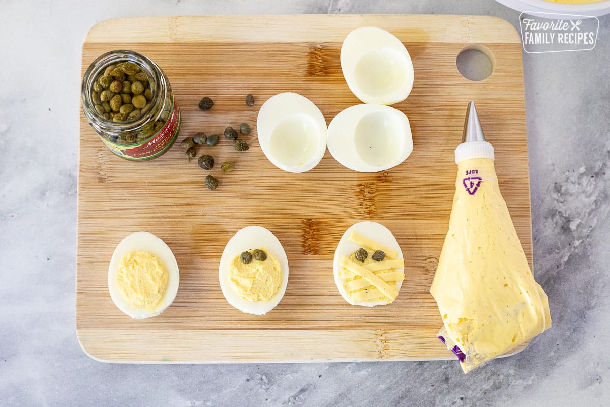 Cutting board with the stages of making a mummy Halloween Deviled Egg with capers and piping bag.