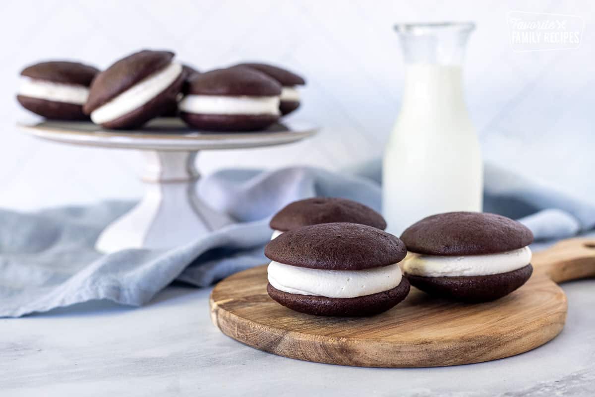 Three Whoopie Pies on a board next to milk and tray of additional whoopie pies.