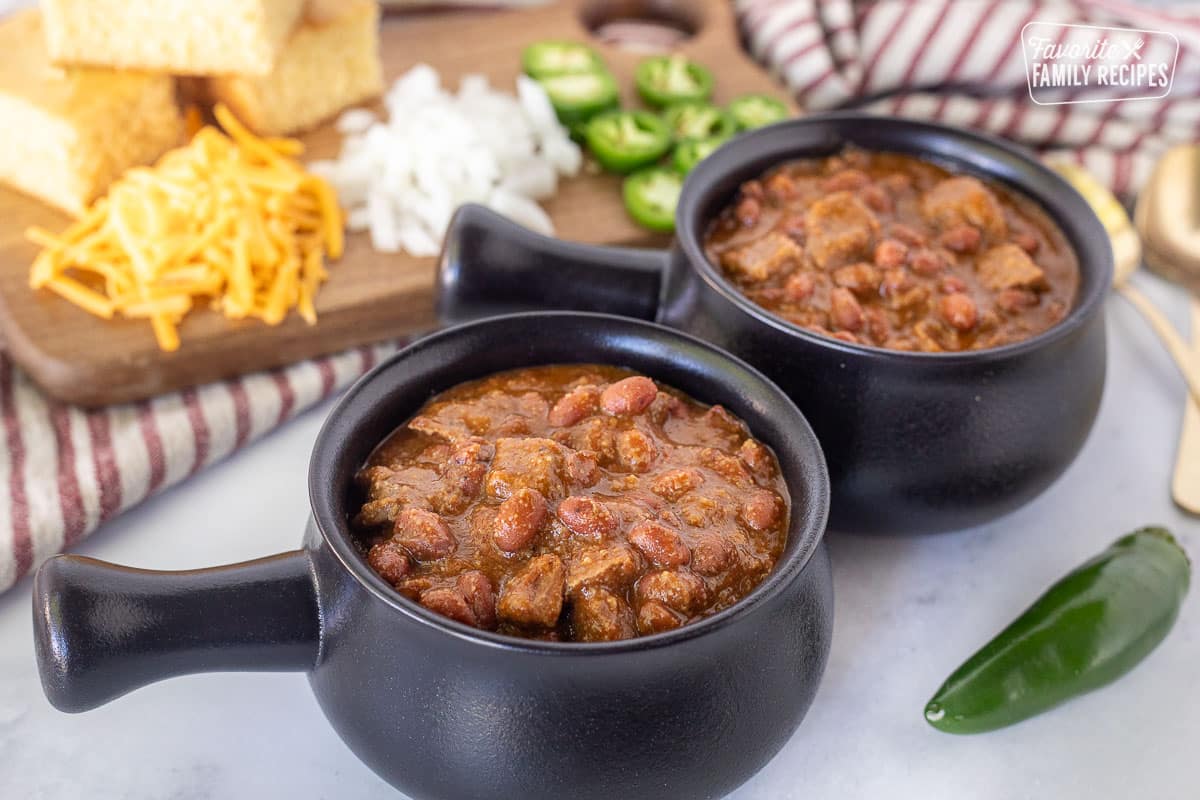 Bowl bowls of Texas Chili without garnish. Cornbread, sliced jalapeño and chopped onions on a cutting board.