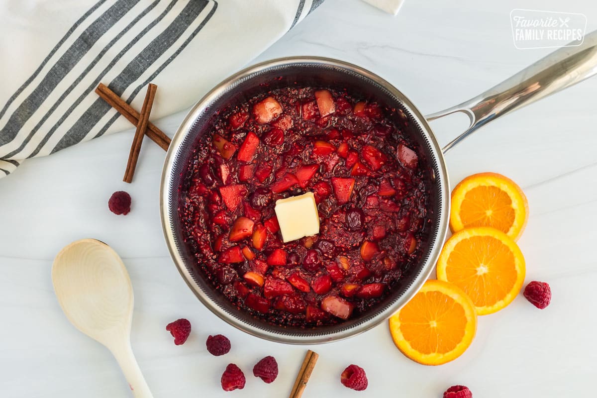 Cooked low sugar cranberry sauce in a saucepan with a small slice of butter on top. Orange slices and raspberries are scattered across table.