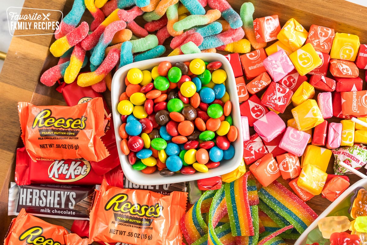 a bowl of M&Ms surrounded by starbursts, sour gummy worms, and chocolates
