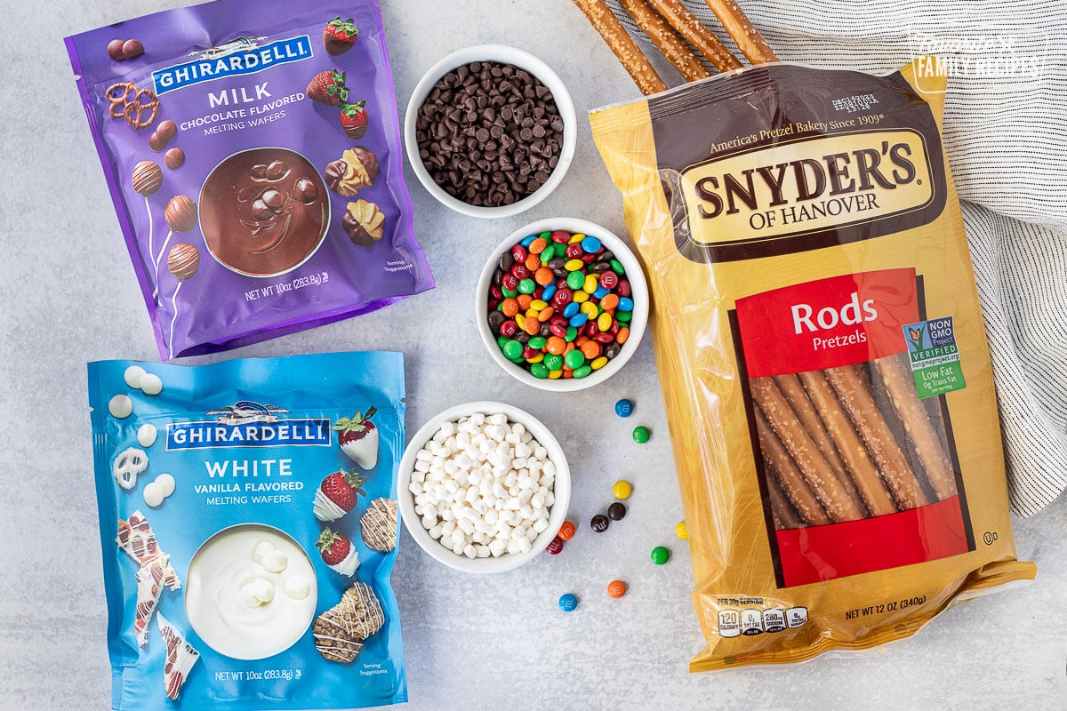Ingredients to make Dipped Pretzel Rods including pretzel rods, Ghirardelli milk chocolate melting wafers, Ghirardelli white chocolate melting wafers, mini mnm's and marshmallow bits.