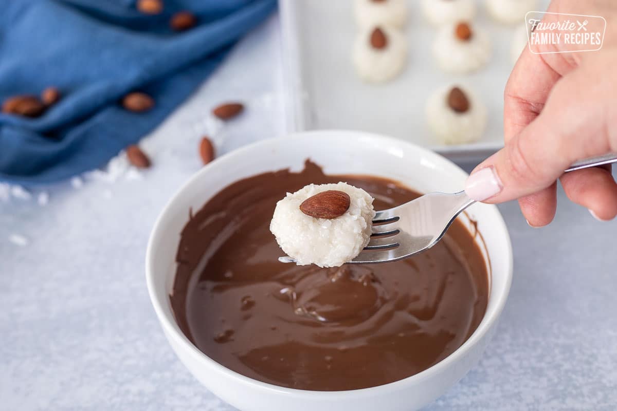 Fork holding a coconut oval ball with an almond on top hovering over a bowl of melted chocolate.
