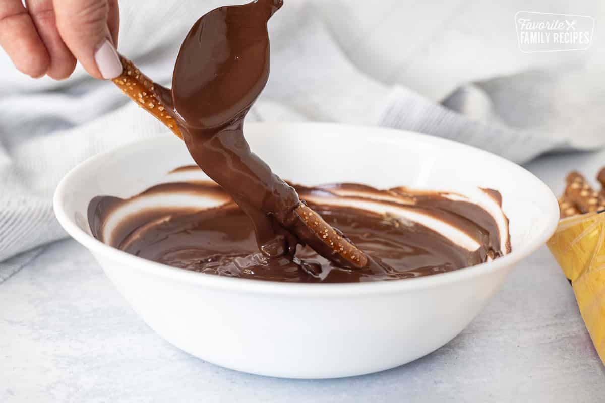 Hand holding pretzel rod into a bowl of melted chocolate with a spoon pouring chocolate onto the pretzel rod.