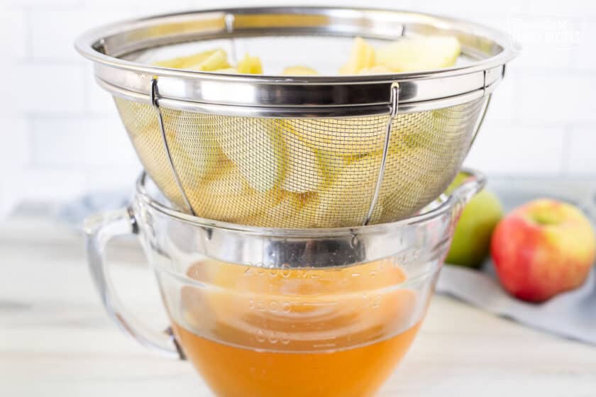 Colander with sliced apples over bowl of apple juice.
