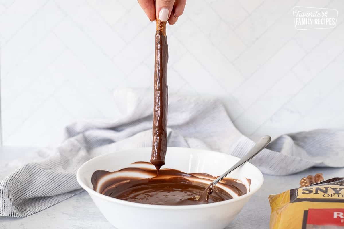 Hand holding a dipped pretzel rod dripping into a bowl of melted chocolate.