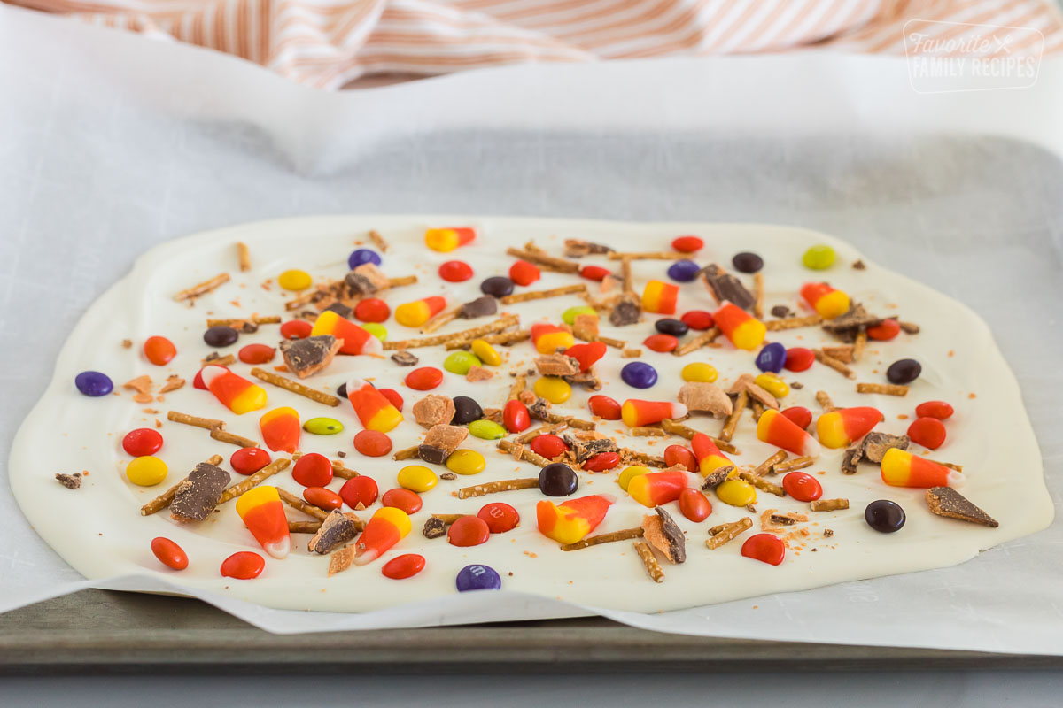 A layer of melted white chocolate beginning to cool on parchment paper on a cookie sheet. It is topped with different candies, including, candy corn, pretzels, m&m's, reese's pieces, and butterfingers.