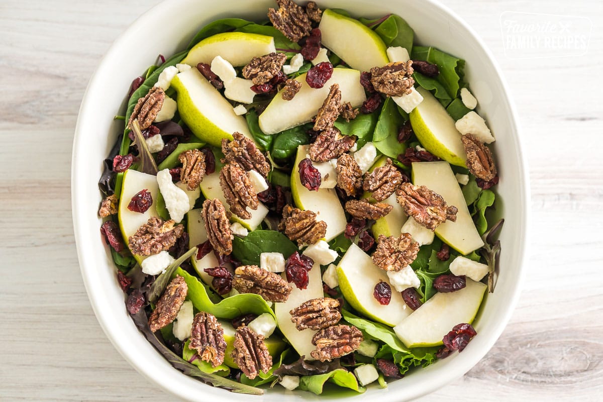 Spring mix in a large bowl with sliced pears, pecans, feta cheese, and dried cranberries