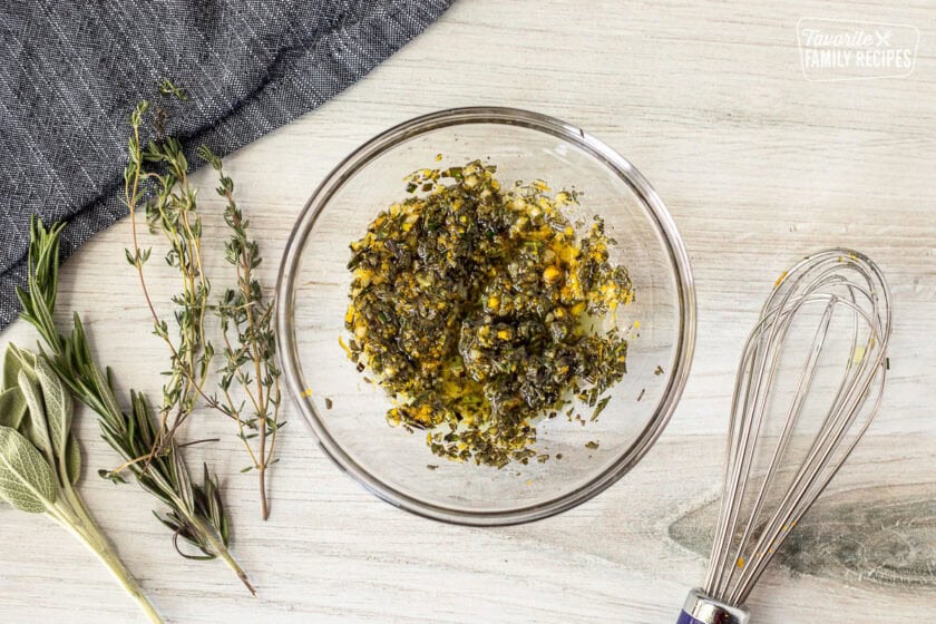 Mixing bowl of herbs and oil. Whisk, sage, rosemary and thyme on the side.