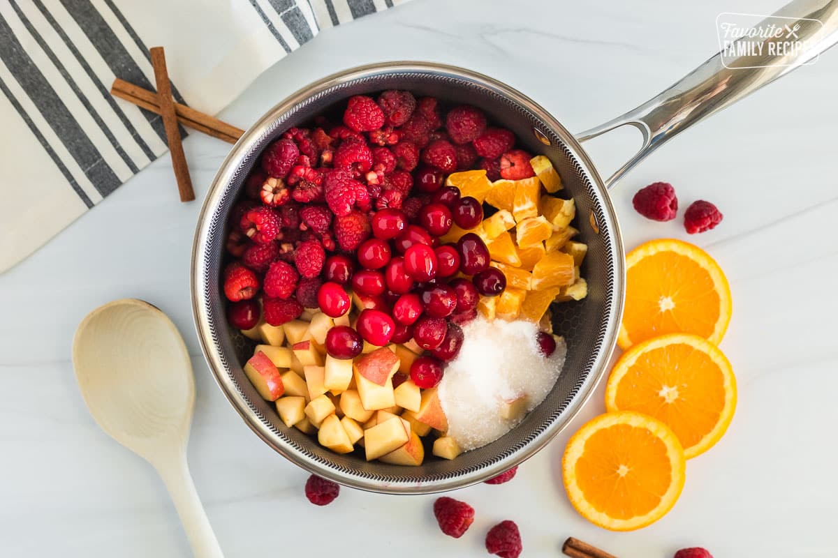 Ingredients for Low Sugar Cranberry Sauce are sitting inside a sauce pan including chopped apples, chopped oranges, cranberries, raspberries, truvia, lemon juice, and maple syrup.