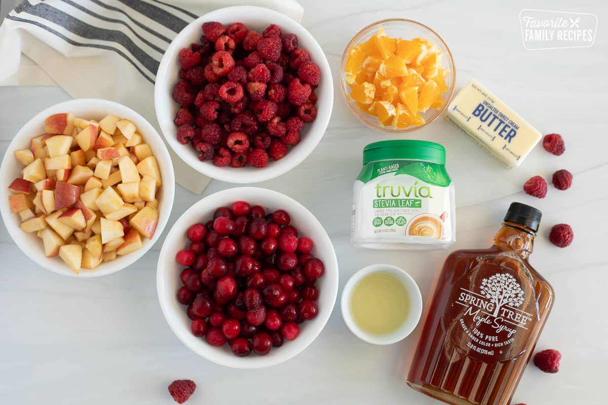 Ingredients for Low Sugar Cranberry sauce are laid out on the table. This includes chopped apples and oranges, cranberries, raspberries, lemon juice, maple syrup, butter, and truvia.