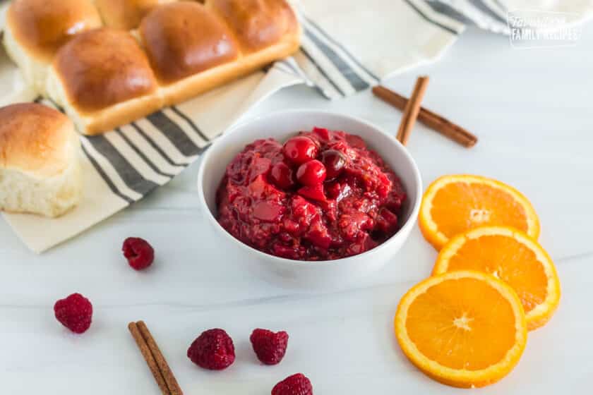 Side view of a bowl containing Low Sugar Cranberry Sauce. Orange slices, cinnamon sticks, raspberries and rolls are also seen scattered across the table.