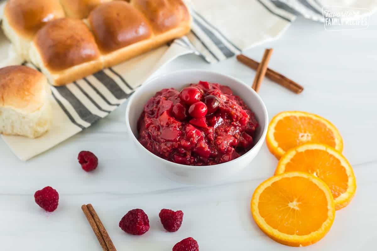Side view of a bowl containing Low Sugar Cranberry Sauce. Orange slices, cinnamon sticks, raspberries and rolls are also seen scattered across the table.