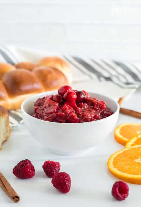Vertical view of a bowl containing Low Sugar Cranberry Sauce. Orange slices, cinnamon sticks, raspberries and rolls are also seen scattered across the table.