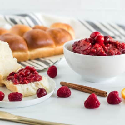 Side view of a bowl of Low Sugar Cranberry sauce, a plate, a knife and rolls. On the plate there is a roll cut open with cranberry sauce spread on top.