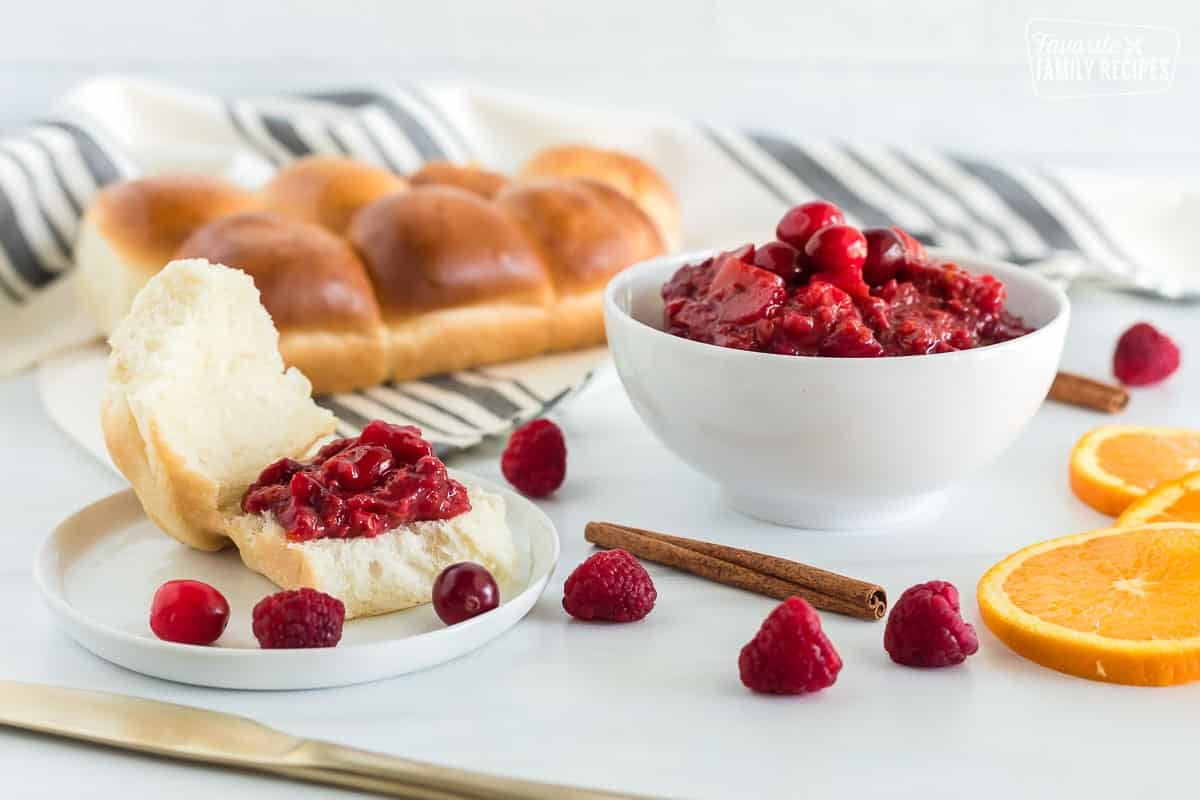 Side view of a bowl of Low Sugar Cranberry sauce, a plate, a knife and rolls. On the plate there is a roll cut open with cranberry sauce spread on top.