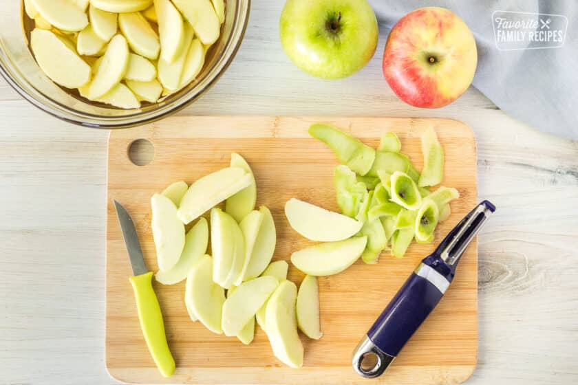 Cutting board with sliced and peeled apples next to a bowl of apples in apple juice.