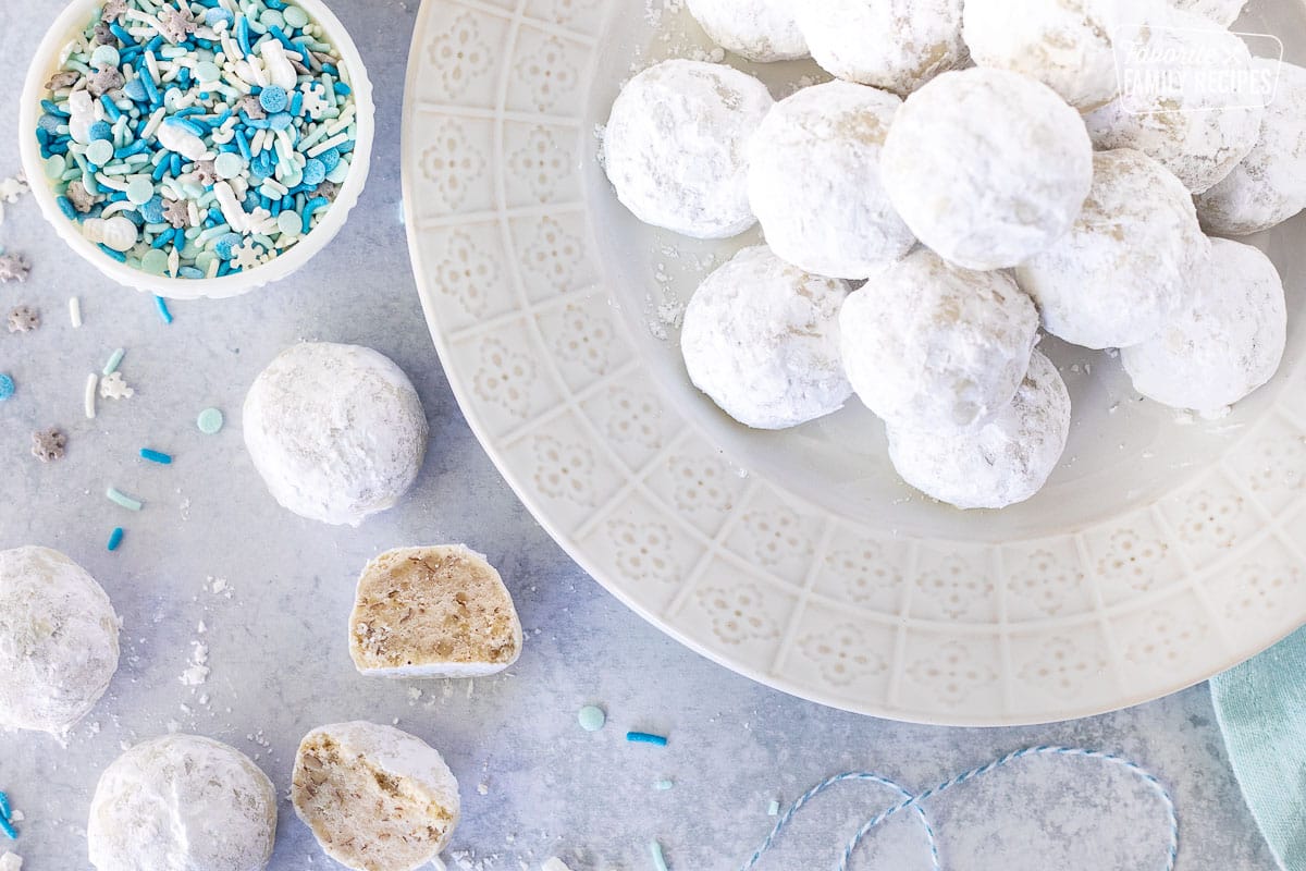 Snowball cookies on a plate next to bowl of sprinkles and cut snowball cookie.