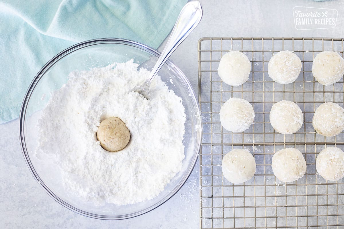 Dipping warm Snowball Cookies into a bowl of powdered sugar next to cooling rack of dipped Snowball cookies.