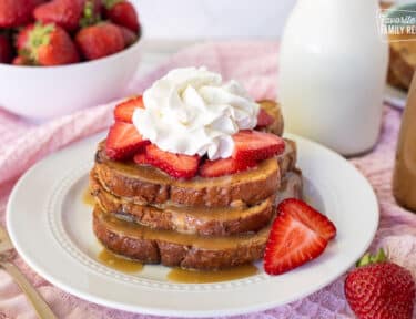 Plate with three slices of Cinnamon French Toast topped with caramel syrup, sliced strawberries and whipped cream.