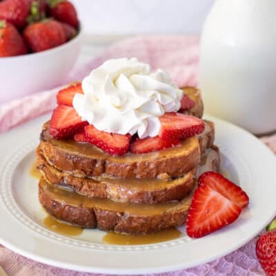 Plate with three slices of Cinnamon French Toast topped with caramel syrup, sliced strawberries and whipped cream.