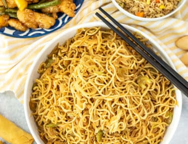 Bowl of Panda Express Chow Mein Noodles next to fried rice, chicken bean, spring rolls and fortune cookies. Chop sticks resting on top of the bowl.