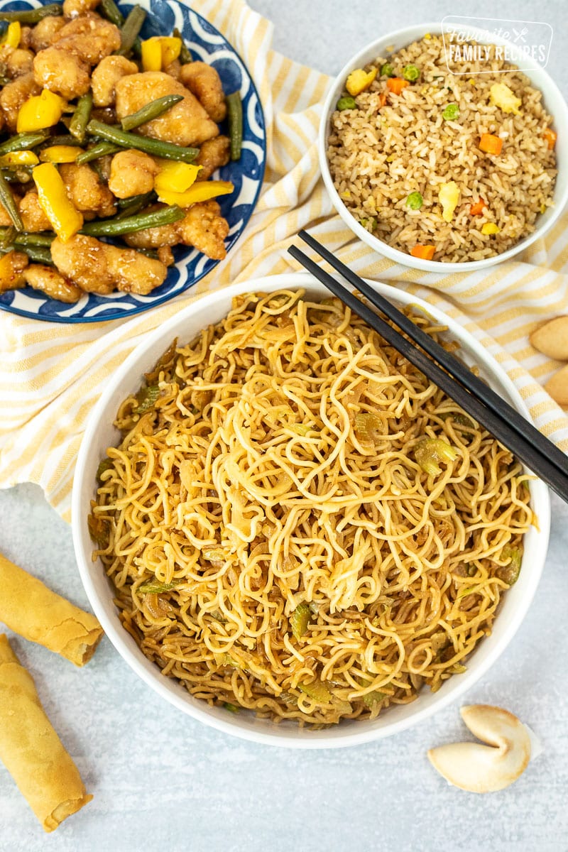 Bowl of Panda Express Chow Mein Noodles next to fried rice, chicken bean, spring rolls and fortune cookies. Chop sticks resting on top of the bowl.