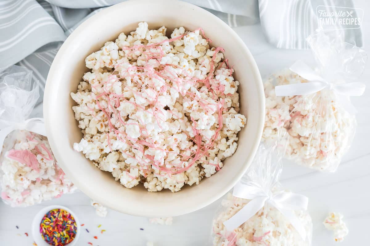 An overhead view of a bowl and three bags containing white chocolate popcorn with ribbons of pink colored chocolate scattered across.
