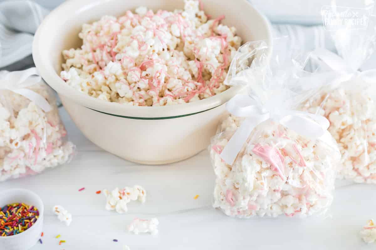 A side view of a bowl and three bags containing white chocolate popcorn with ribbons of pink colored chocolate scattered across.