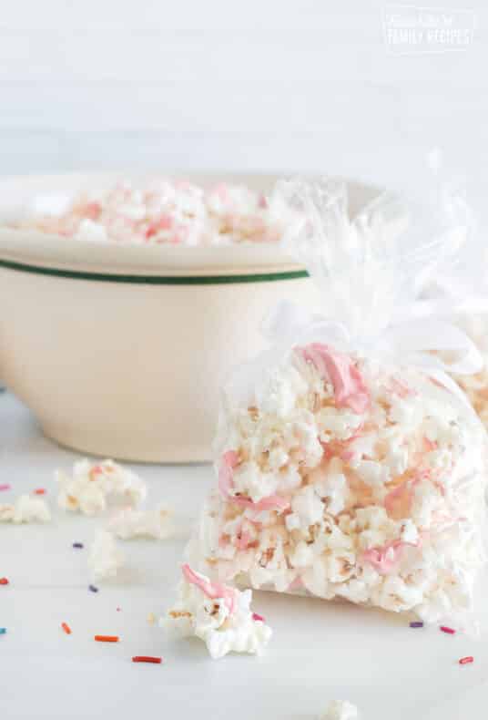 Vertical view of bags and a bowl of white chocolate popcorn. The popcorn has ribbons of melted pink chocolate across the pieces.