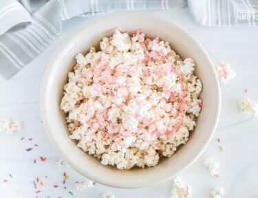 An overhead view of a bowl containing white chocolate popcorn with ribbons of pink colored chocolate scattered across.