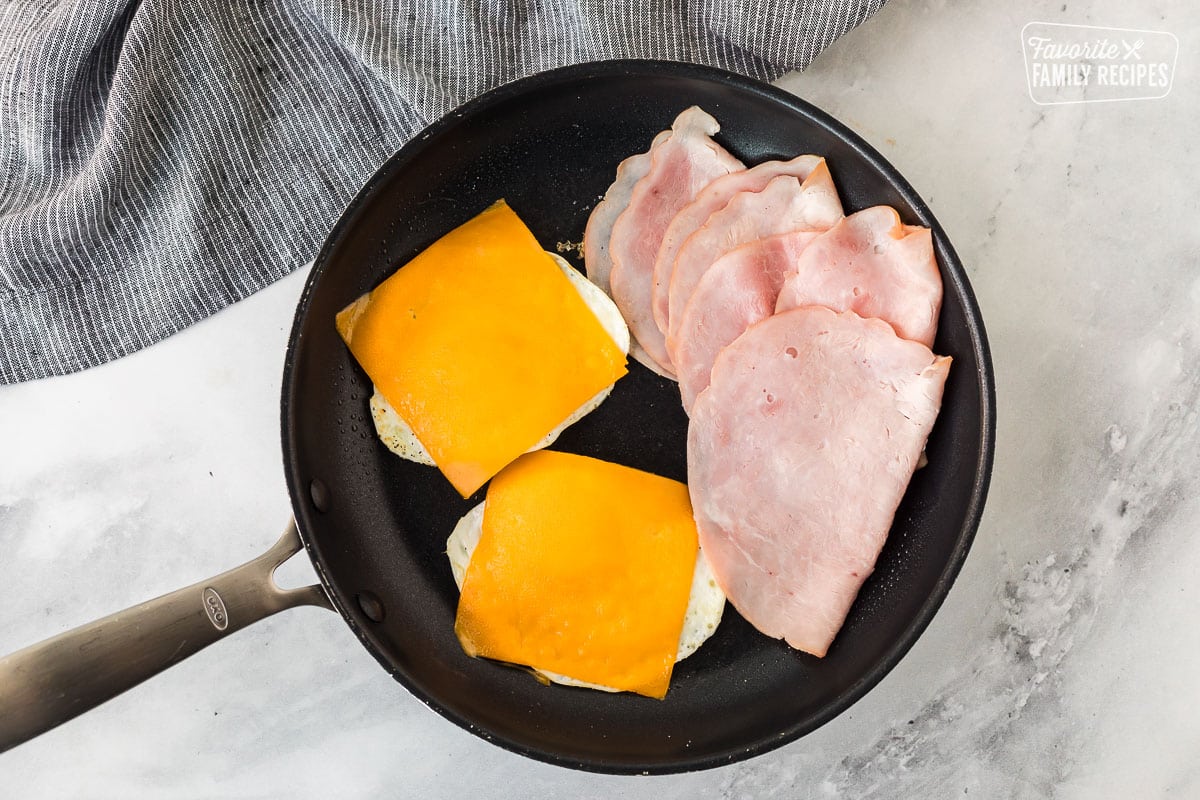 Skillet with two eggs covered in cheese slices and ham on the side.