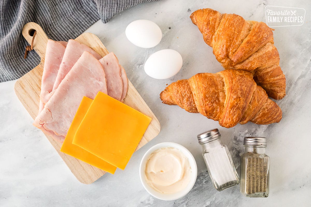 Ingredients to make a Croissant Breakfast Sandwich including two croissants, sliced cheddar cheese, sliced black forrest ham, eggs, mayo, salt and pepper.